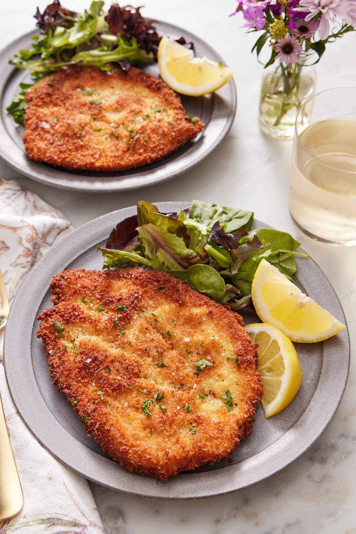 A plate with a crispy chicken schnitzel, mixed greens, and lemon wedges. A drink, vase of flowers, and another plated serving of chicken schnitzel in the back.