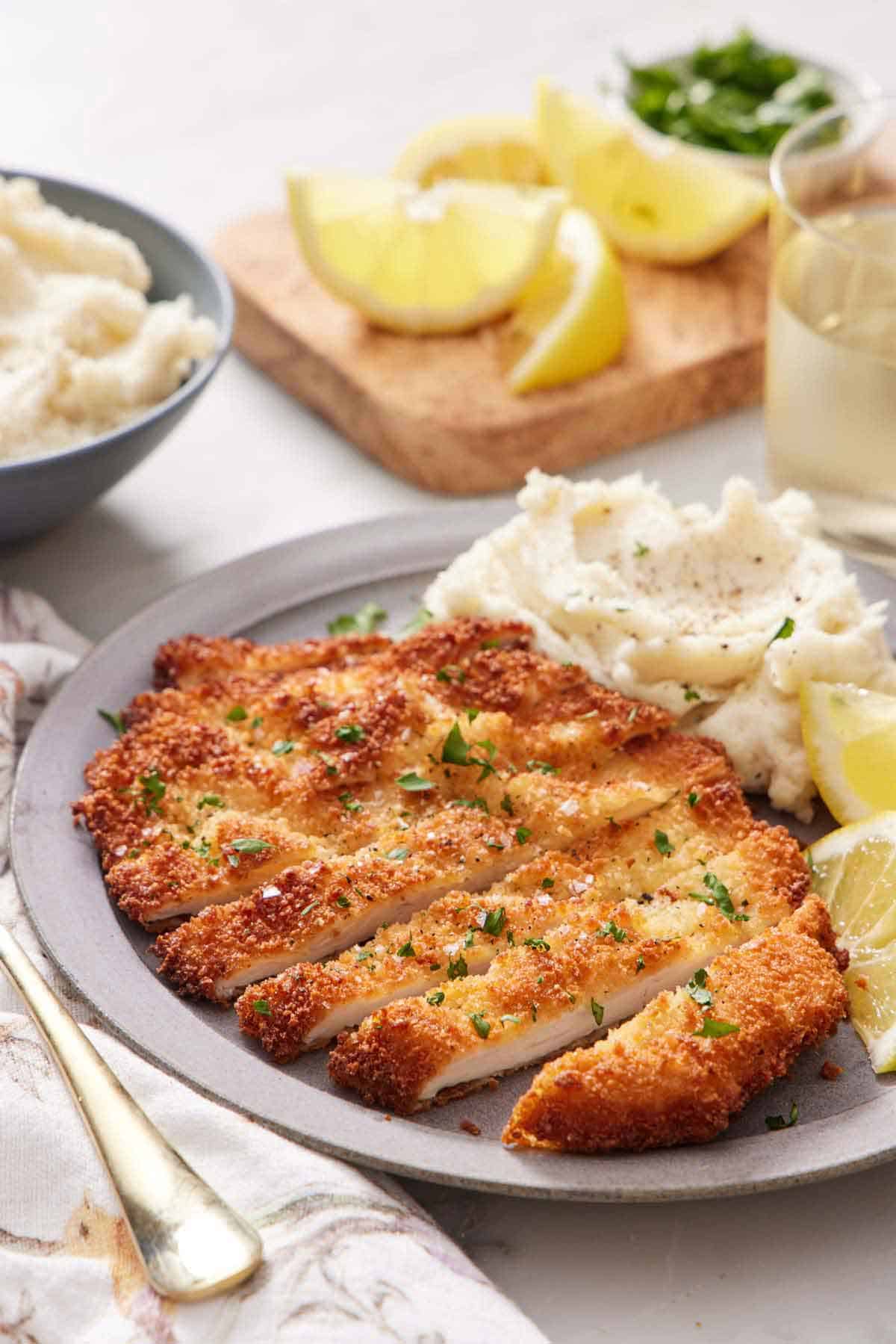 A sliced chicken schnitzel on a plate with lemon wedges and mashed potatoes. A drink in the background along with cut lemons and bowl of mashed potatoes.