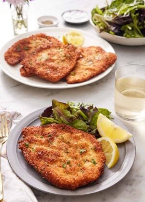A plate with a crispy chicken schnitzel, mixed greens, and lemon wedges. A drink in the back along with a platter with more chicken schnitzels and a bowl of mixed greens.