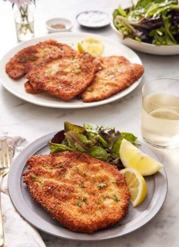 A plate with a crispy chicken schnitzel, mixed greens, and lemon wedges. A drink in the back along with a platter with more chicken schnitzels and a bowl of mixed greens.