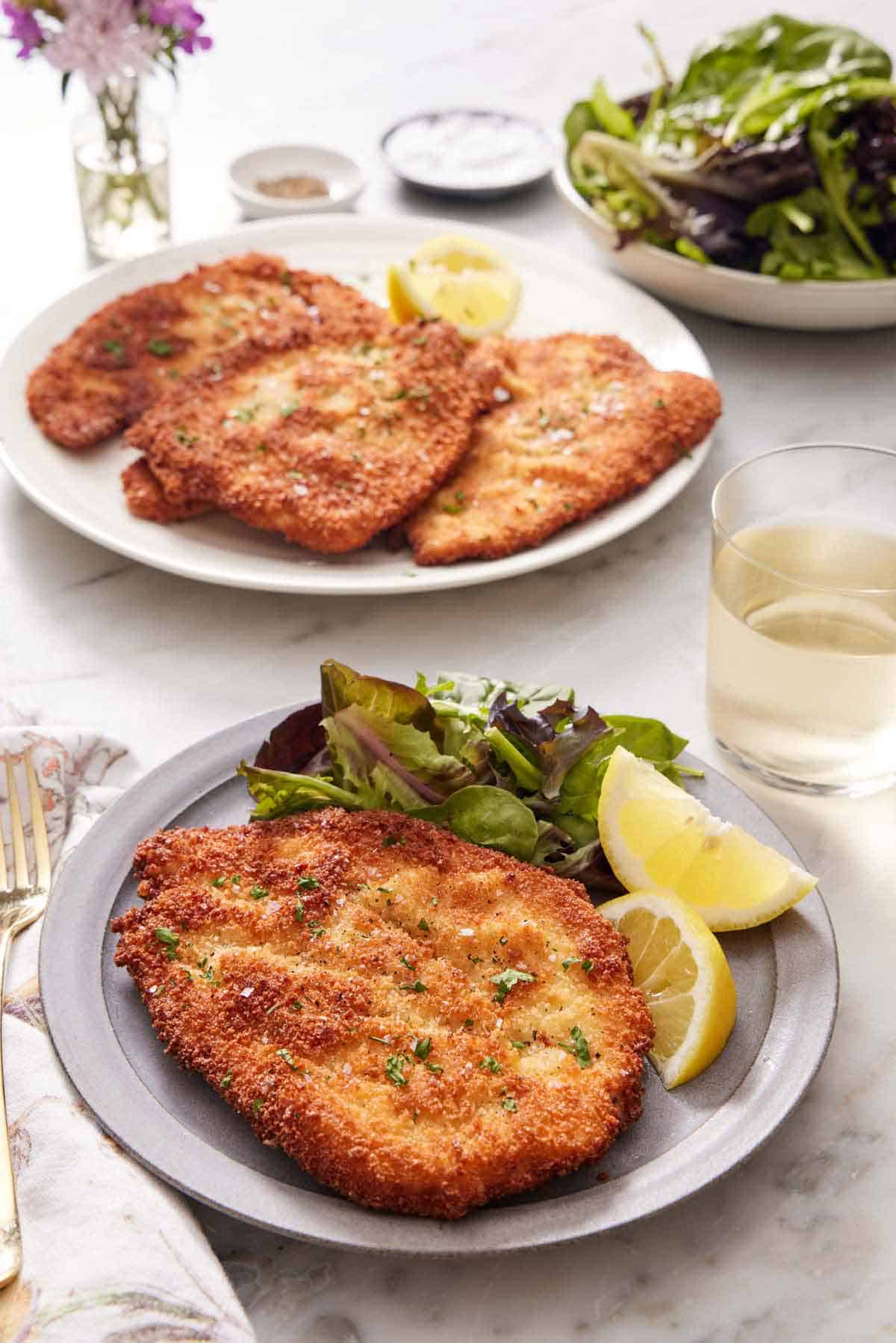 A plate with a crispy chicken schnitzel, mixed greens, and lemon wedges. A drink in the back along with a platter with more chicken schnitzels and a bowl of mixed greens.