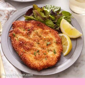 Pinterest graphic of a plate with a crispy chicken schnitzel, mixed greens, and lemon wedges.