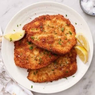 Overhead view of a platter of chicken schnitzels with lemon wedges along garnished with chopped parsley. A bowl of salt on the side and linen napkin.