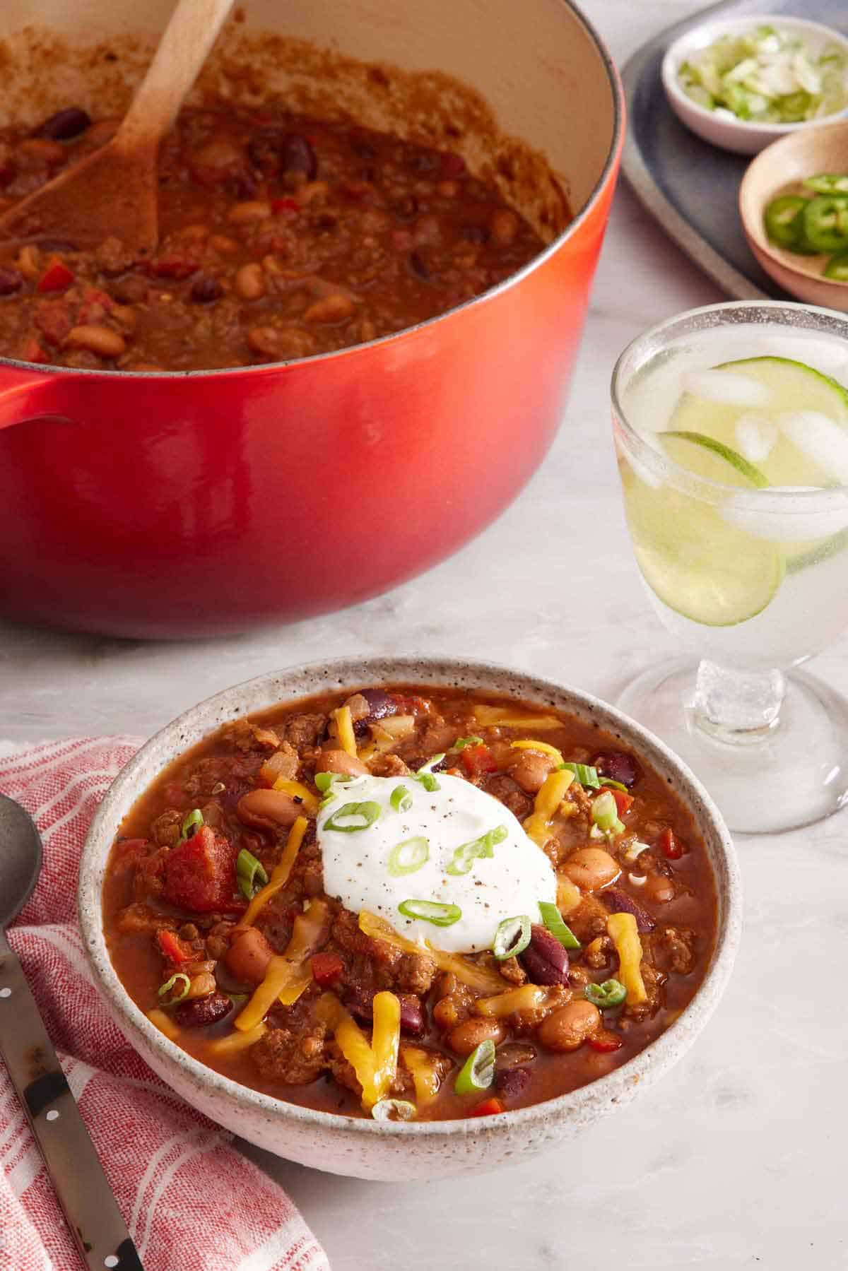 A bowl of chili topped with sour cream, green onions, and shredded cheese. A drink with limes in the back along with a dutch oven of more chili.