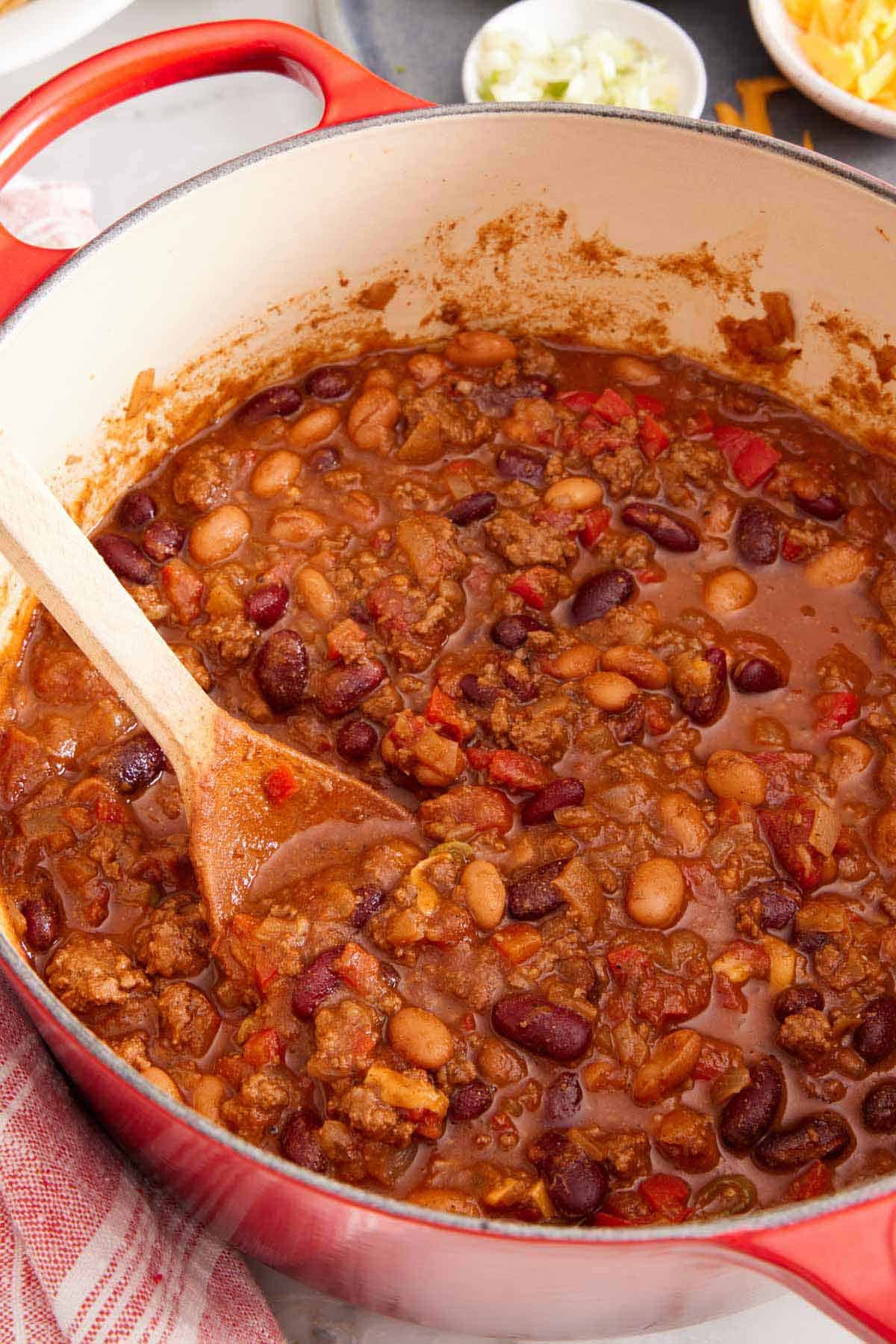 A red dutch oven of chili with a wooden spoon inside.