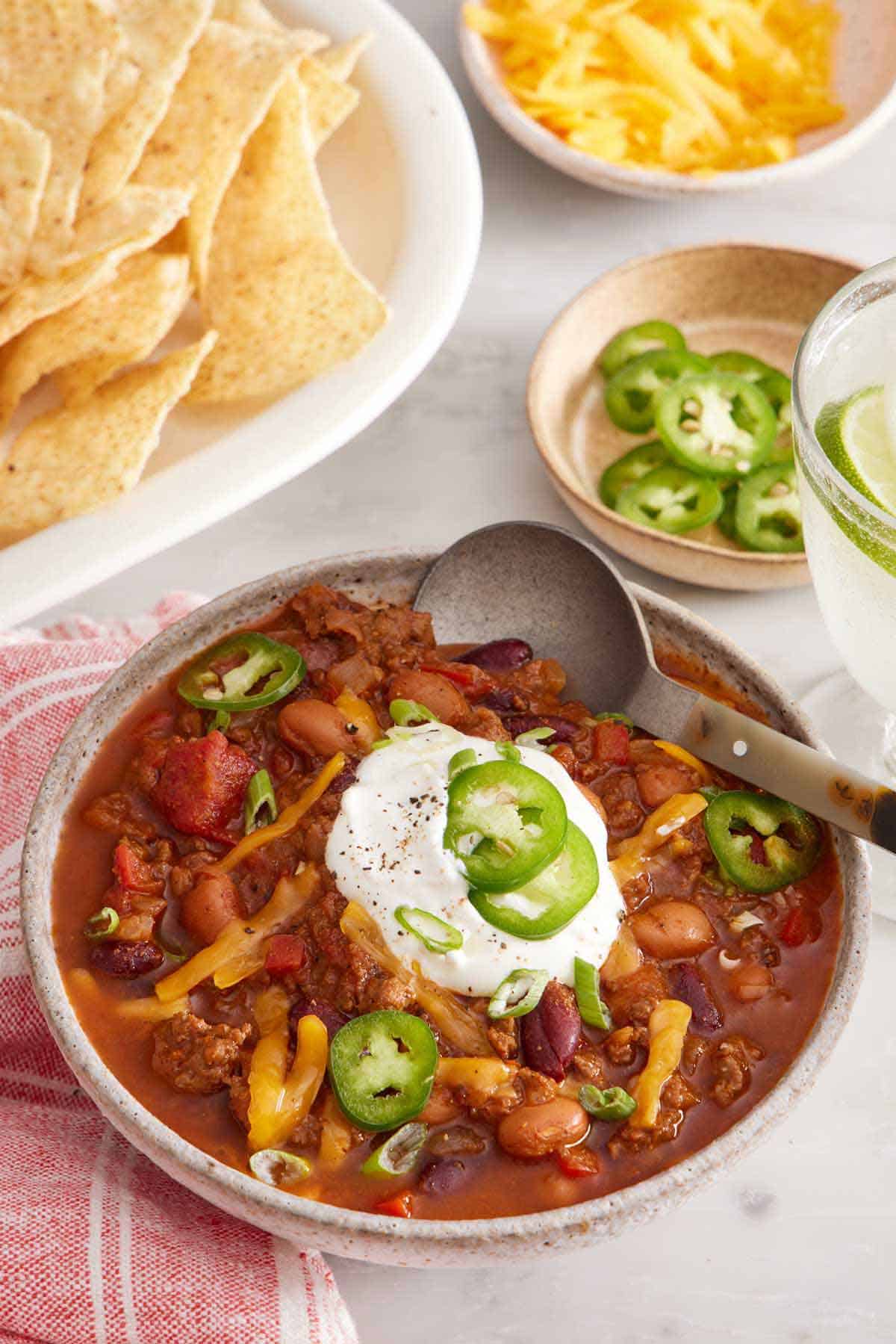 A bowl of chili with sliced jalapeno, sour cream, and shredded cheese with a spoon tucked in. Tortilla chips, jalapenos, and shredded cheese in the background.