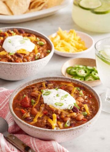 A bowl of chili topped with sour cream, shredded cheese, and green onions. Another bowl in the background along with bowls of toppings.