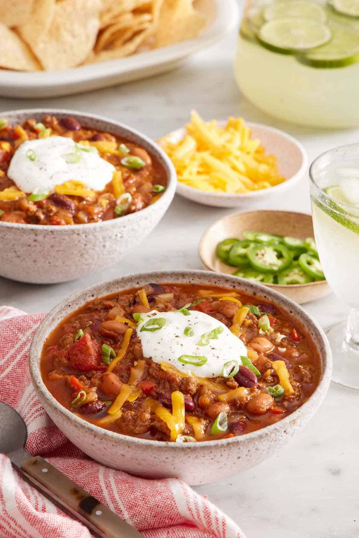 A bowl of chili topped with sour cream, shredded cheese, and green onions. Another bowl in the background along with bowls of toppings.