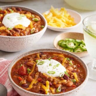 Pinterest graphic of a bowl of chili topped with sour cream, shredded cheese, and green onions. Another bowl in the background along with bowls of toppings.