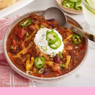 Pinterest graphic of a bowl of chili with sliced jalapeno, sour cream, and shredded cheese with a spoon tucked in.