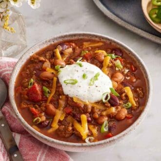 A bowl of chili with a dollop of sour cream, shredded cheese, and green onions on top. A spoon on the side along with a bowl of sliced jalapenos.