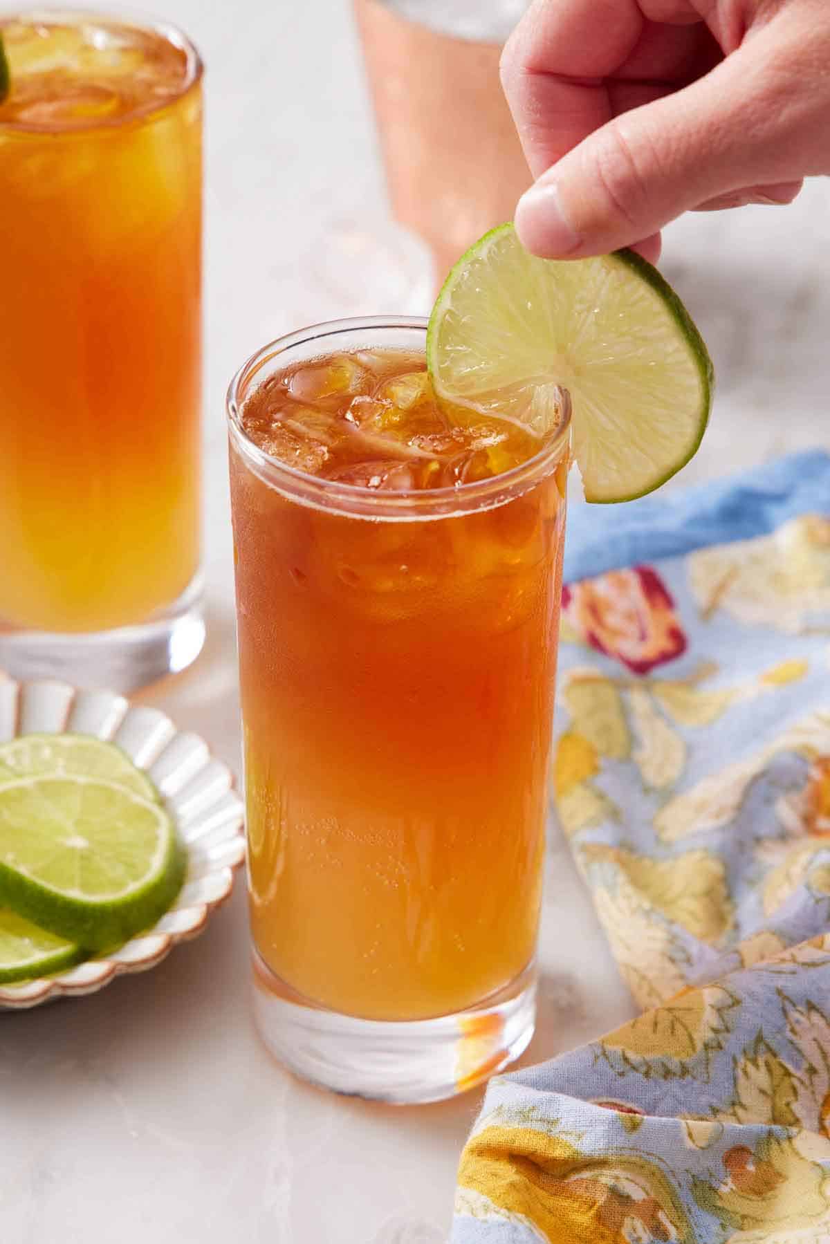 A glass of Dark and Stormy with a lime wheel placed onto the rim. More cut limes and another cocktail in the background.