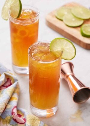 Two glasses of Dark and Stormy cocktails with a lime wheel on the edge. More lime wheels in the background along with a jigger.