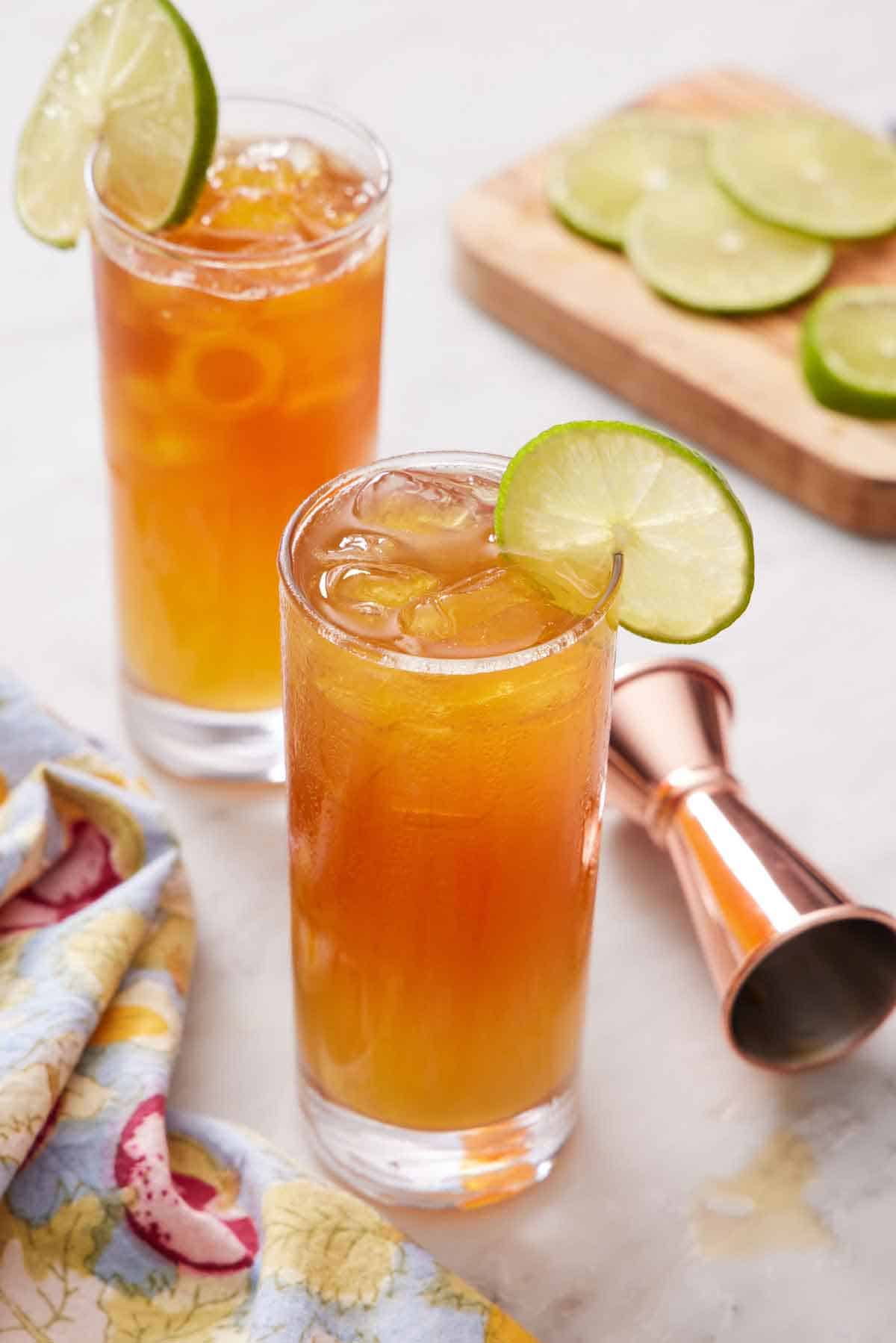 Two glasses of Dark and Stormy cocktails with a lime wheel on the edge. More lime wheels in the background along with a jigger.