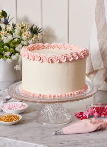 A cake with white frosting and pink frosting piped on the top and bottom of the cake on a cake stand. A piping bag and bowls of sprinkles on the counter.