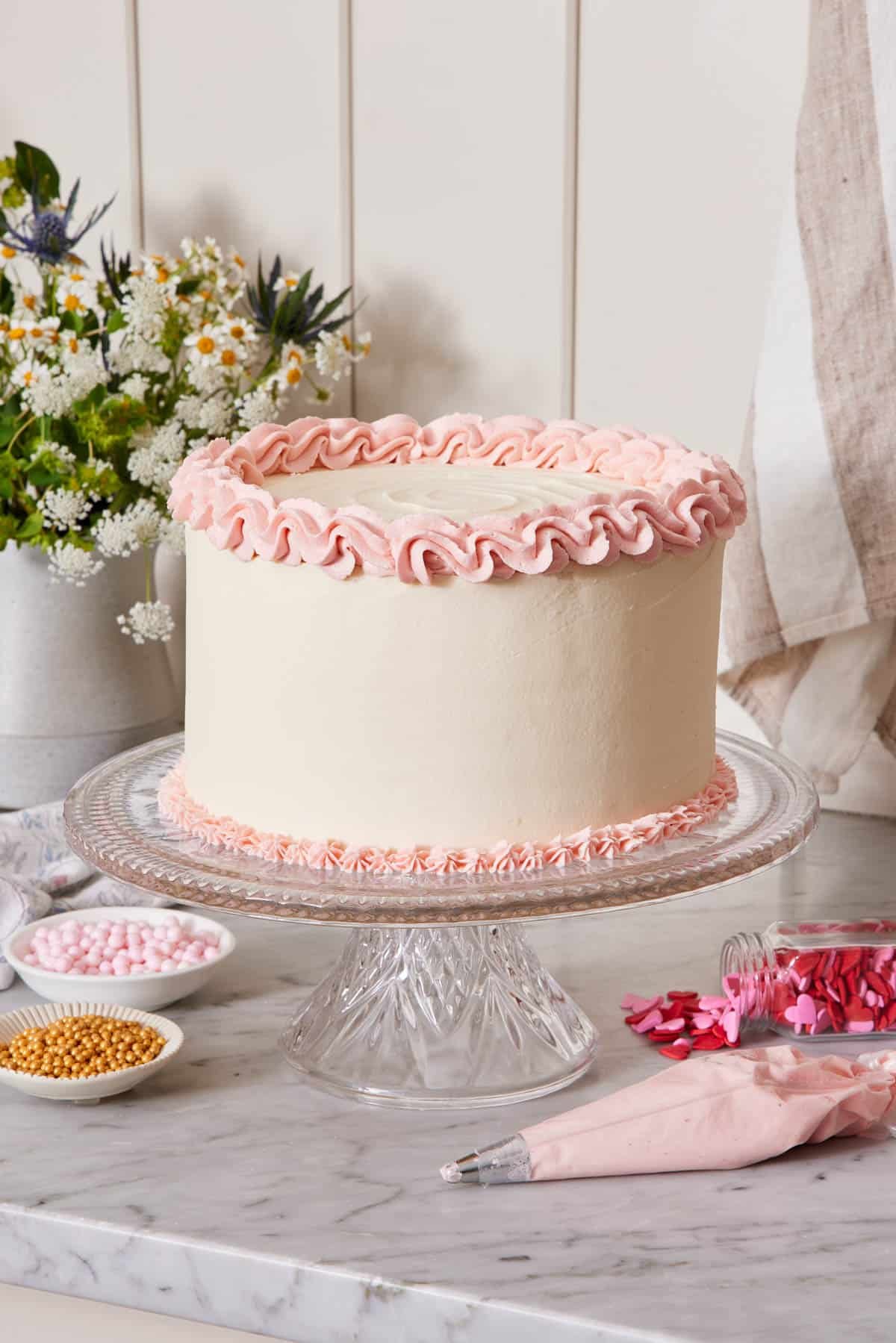 A cake with white frosting and pink frosting piped on the top and bottom of the cake on a cake stand. A piping bag and bowls of sprinkles on the counter.