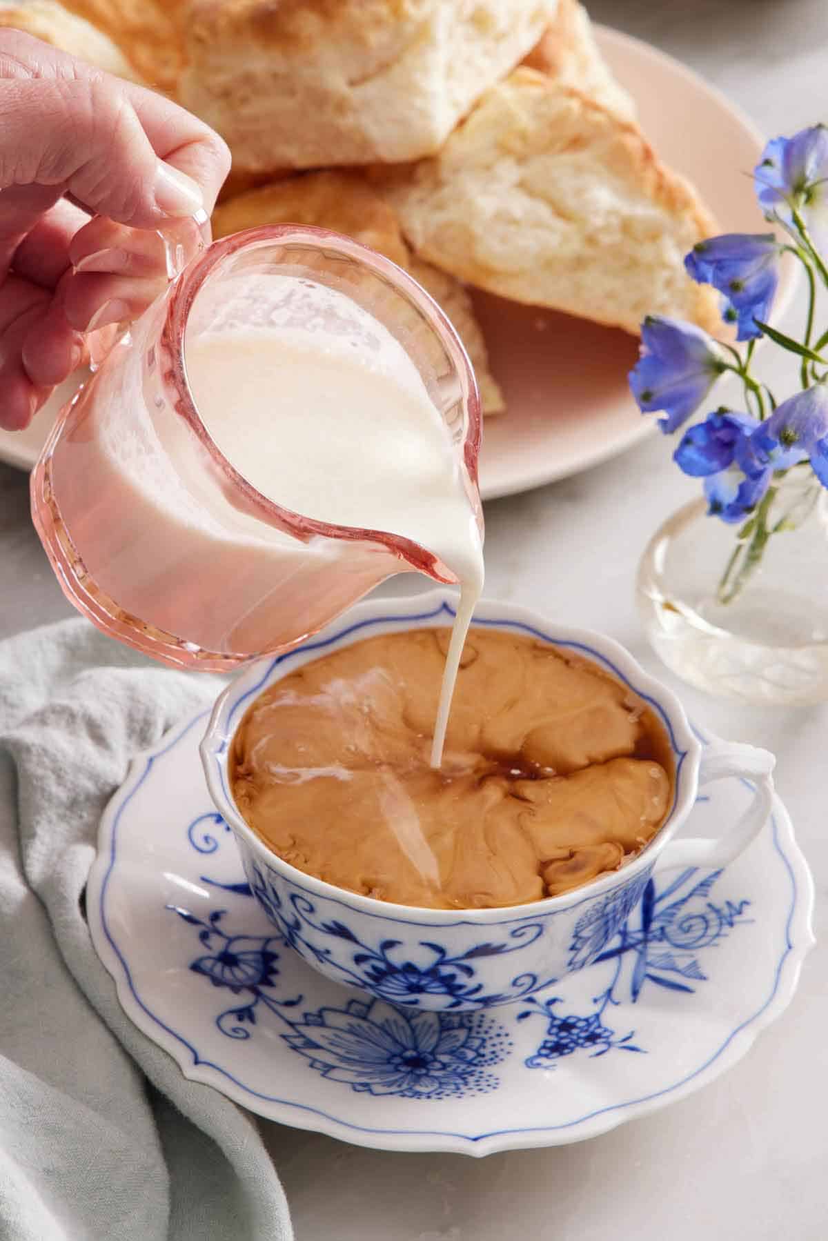 Heavy cream poured into a cup of tea with on a plate. A platter of biscuits in the background.