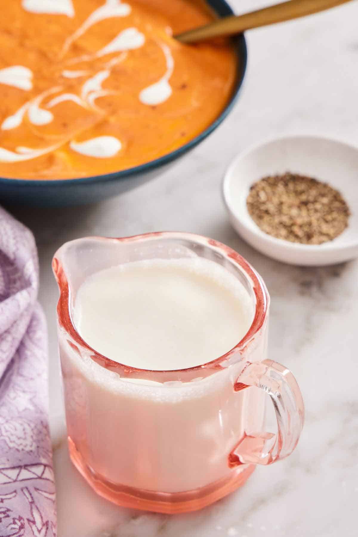 A mini jug with homemade heavy cream. A small bowl of pepper and bowl of soup in the background.