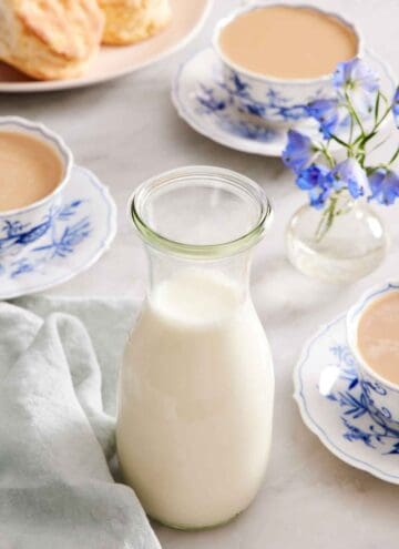 A jar of homemade heavy cream. Three cups of tea with heavy cream in the background along with biscuits and flowers.