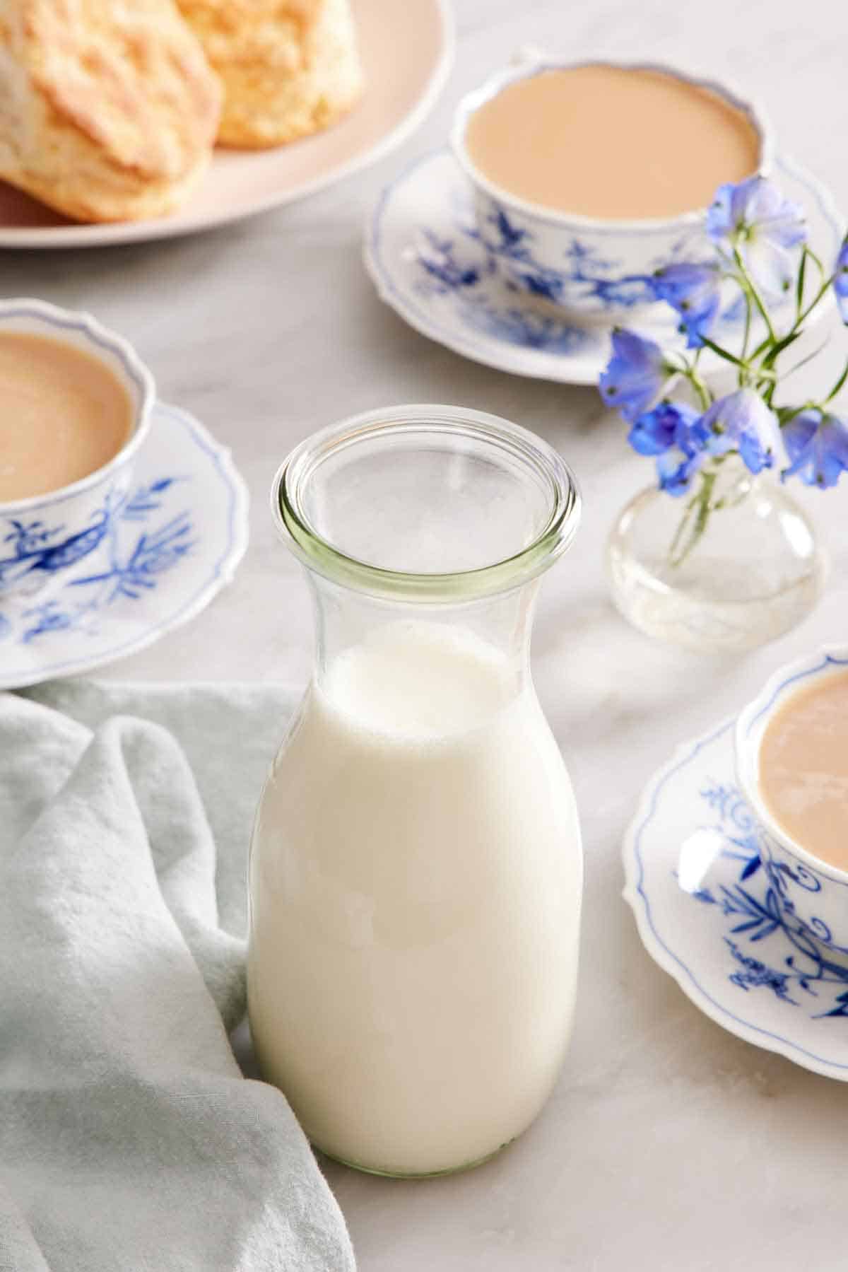 A jar of homemade heavy cream. Three cups of tea with heavy cream in the background along with biscuits and flowers.