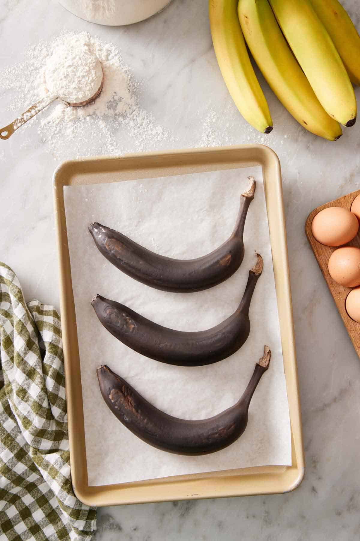 Three oven-ripened bananas on a parchment lined sheet pan on a marble surface. Eggs, bananas, and a scoop of flour off to the side.