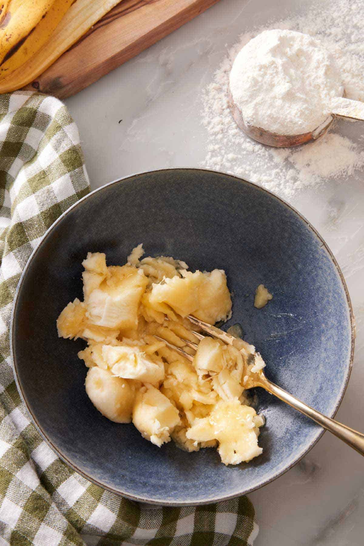 Ripe banana mashed with a fork in a bowl. A scoop of flour in the background.