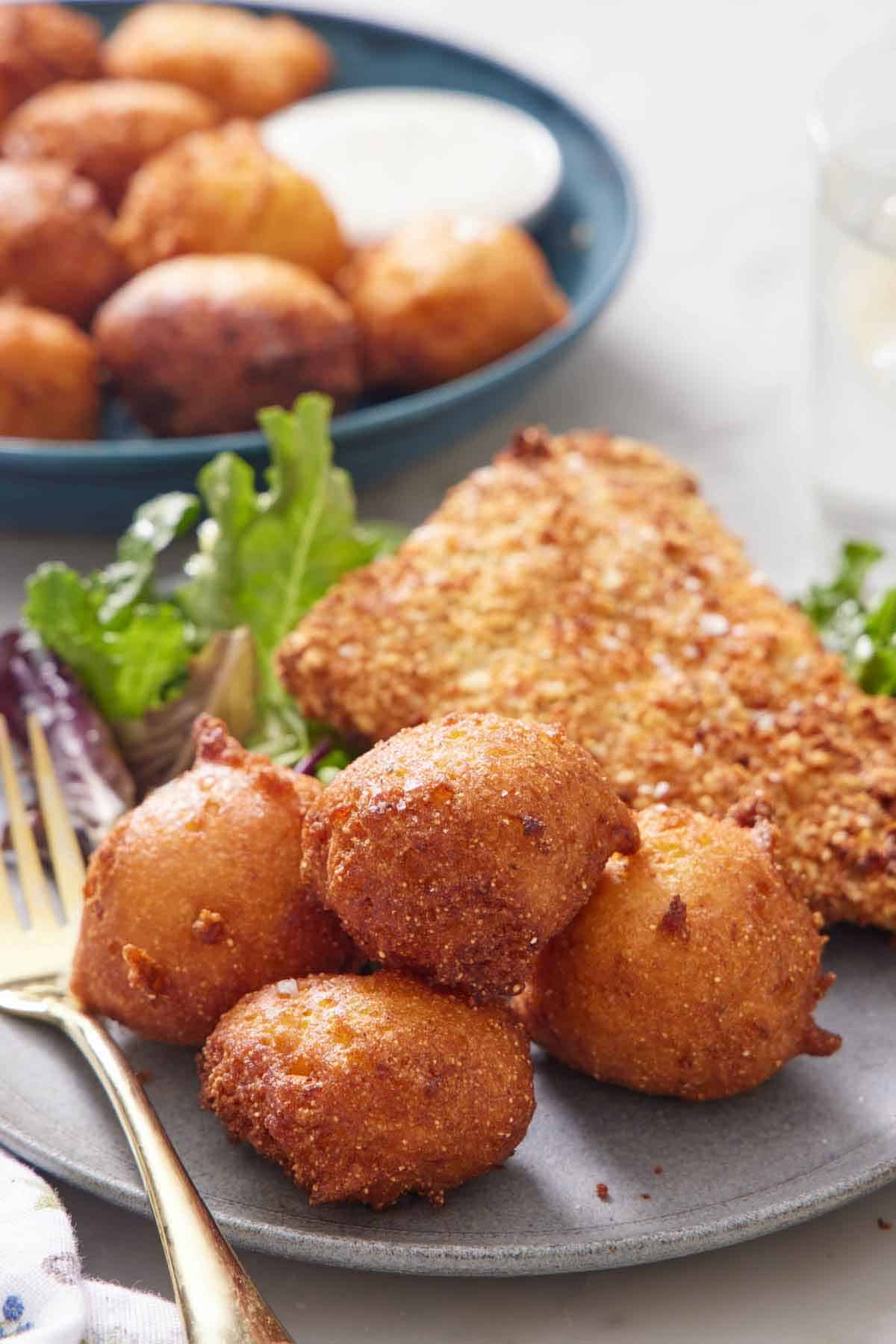 A plate with four hush puppies with fried fish, mixed greens, and a fork. More hush puppies in the background.