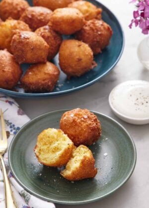 A green plate with two hush puppies, with one torn in half. A large plate of more hush puppies in the background along with a dipping sauce.