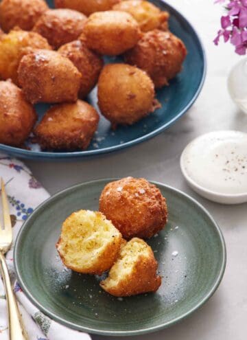 A green plate with two hush puppies, with one torn in half. A large plate of more hush puppies in the background along with a dipping sauce.