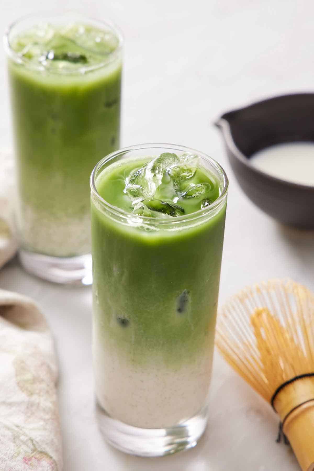 Two glasses of iced matcha lattes with the milk and matcha layers separated. A matcha whisk on the side.