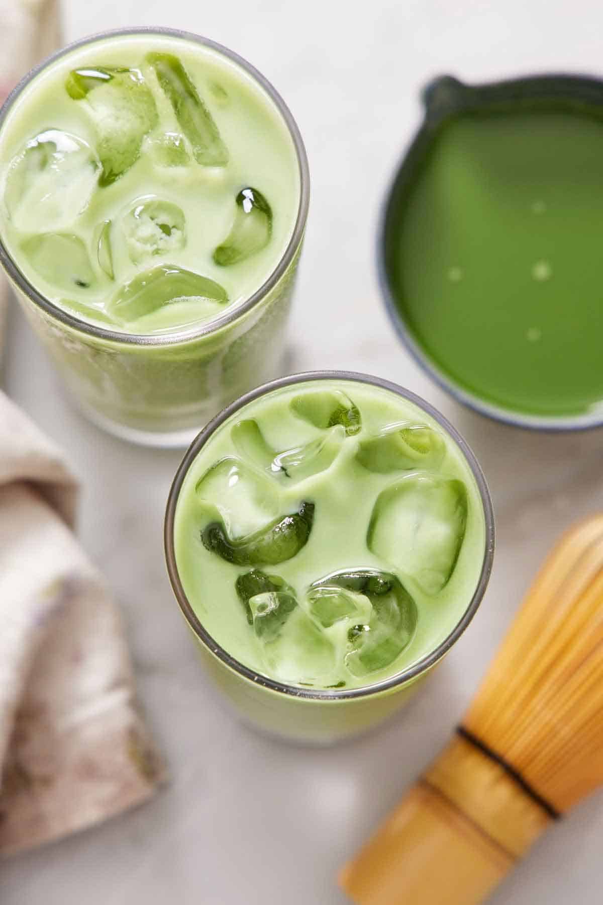 Overhead view of two glasses of iced matcha lattes. A matcha bowl with more matcha and a matcha whisk beside them.