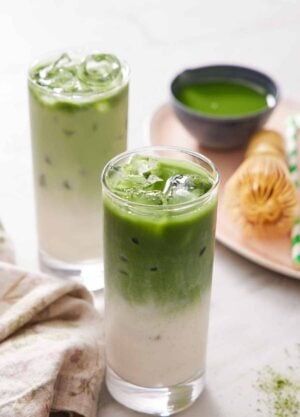 Two glasses of iced matcha lattes. A platter in the background with more matcha, a matcha whisk, and a straw.