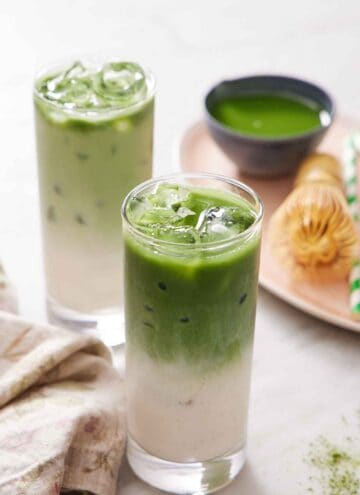 Two glasses of iced matcha lattes. A platter in the background with more matcha, a matcha whisk, and a straw.