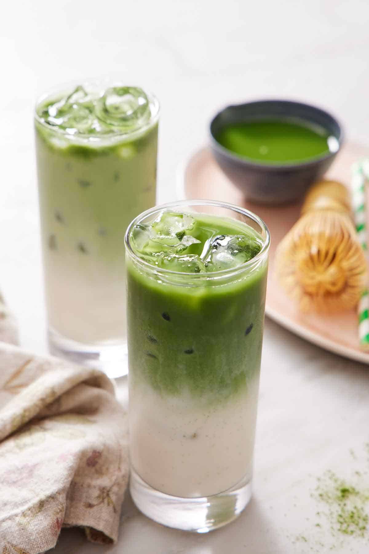 Two glasses of iced matcha lattes. A platter in the background with more matcha, a matcha whisk, and a straw.