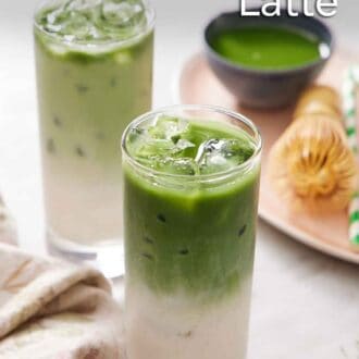 Pinterest graphic of two glasses of iced matcha latte. A platter in the background with more matcha, a matcha whisk, and a straw.