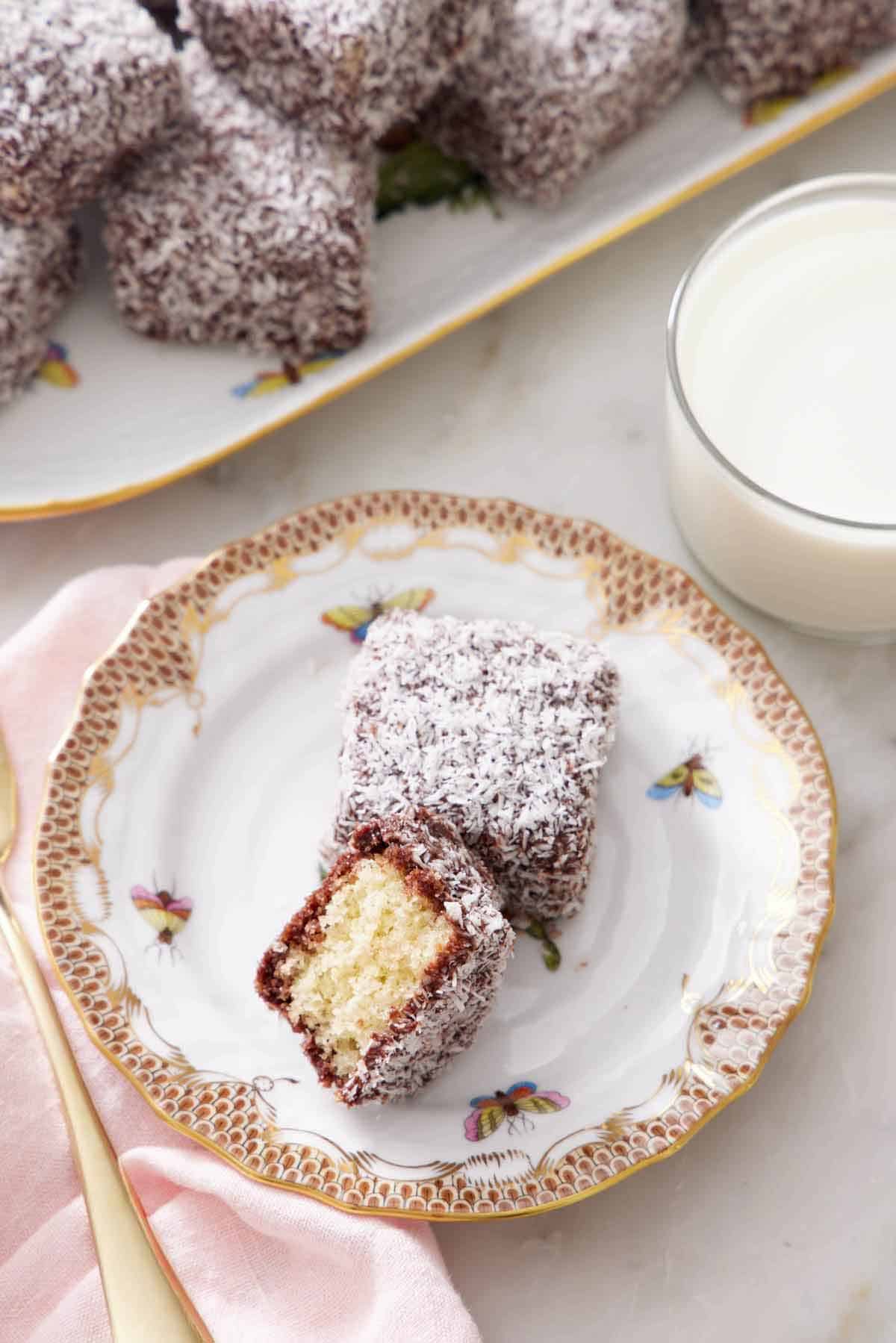 A plate with a lamington and a half with the interior facing up. A glass of milk and more lamingtons off to the side.