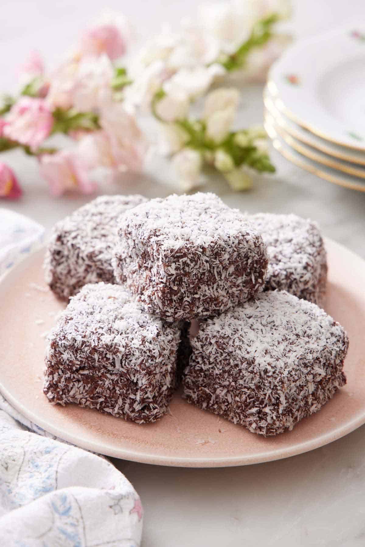 A pink plate with a small pile of five lamingtons. Flowers in the background along with a stack of plates.