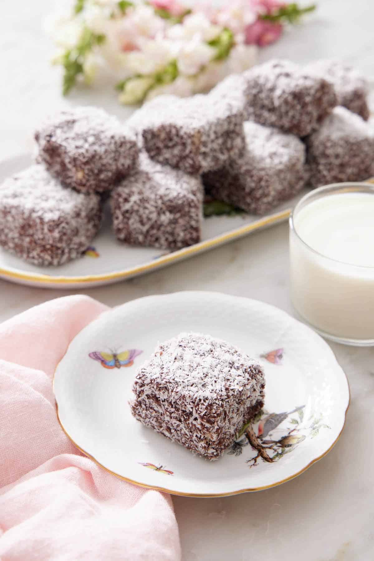 A plate with a lamington with a glass of milk and platter of more lamingtons.