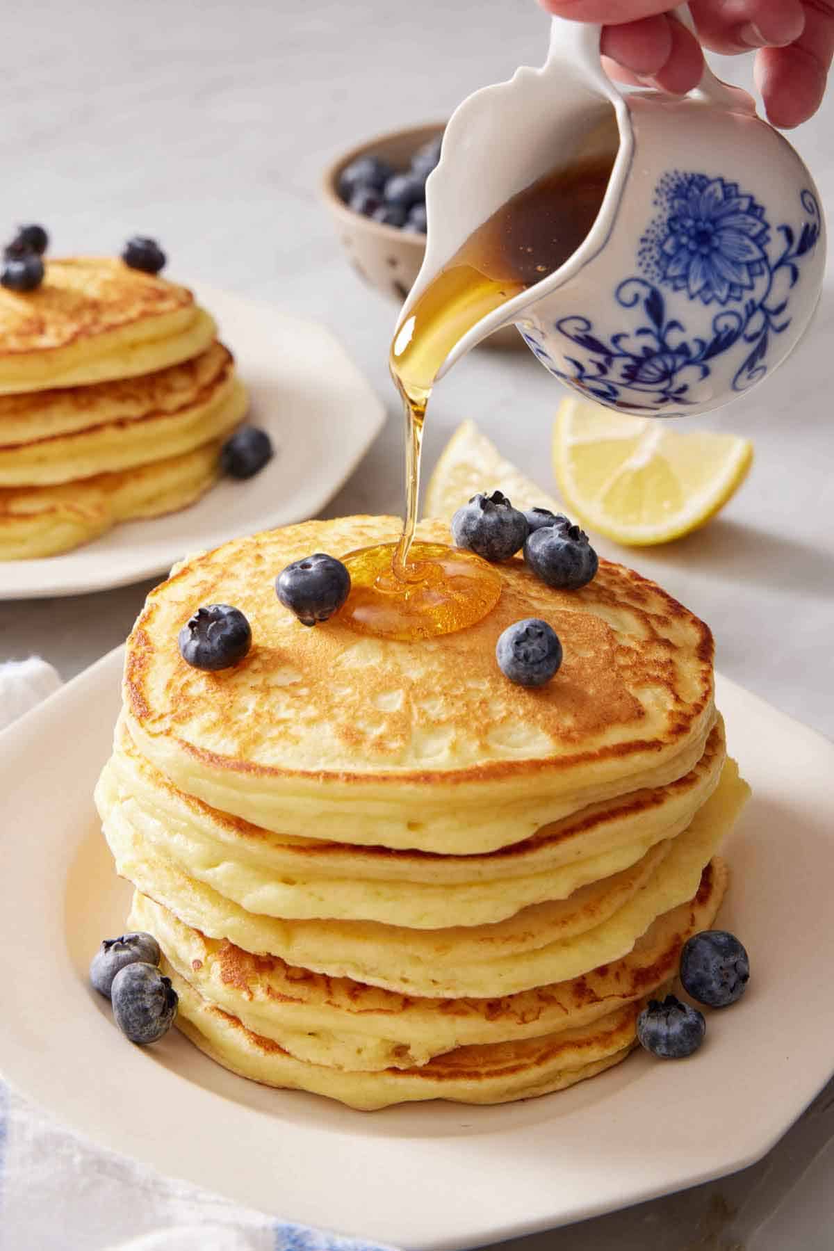 Maple syrup poured over a stack of lemon ricotta pancakes with fresh blueberries. Another stack of pancakes in the background along with some lemon wedges and blueberries.