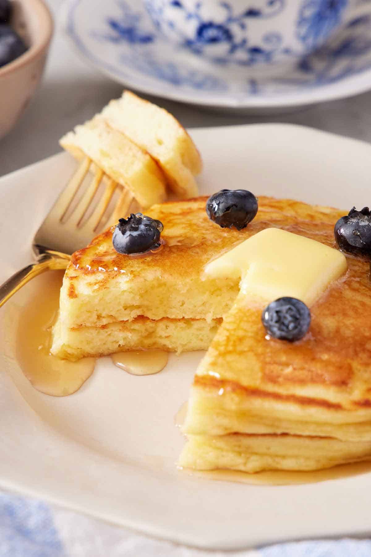 A plate with two lemon ricotta pancakes with butter, maple syrup, and fresh blueberries on top. A bite cut out and on a fork on the side.