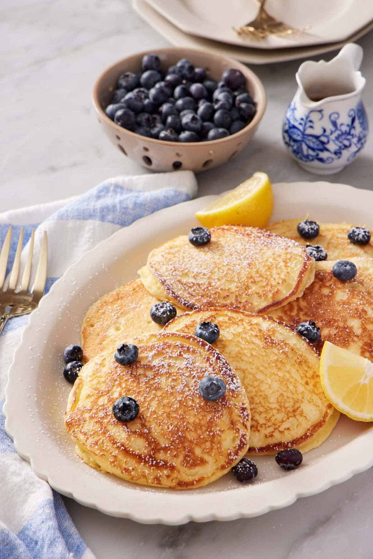 A platter of lemon ricotta pancakes with fresh blueberries and lemon wedges. A dusting of powdered sugar on top. A bowl of blueberries and jug of maple syrup in the background.