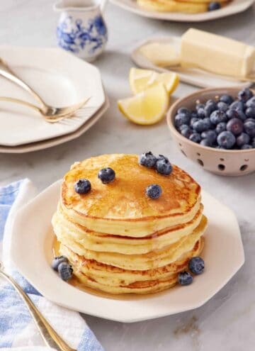 A stack of lemon ricotta pancakes topped with maple syrup and blueberries. A bowl of blueberries in the background along with plates, forks, lemon wedges, butter, and a small jug of syrup.