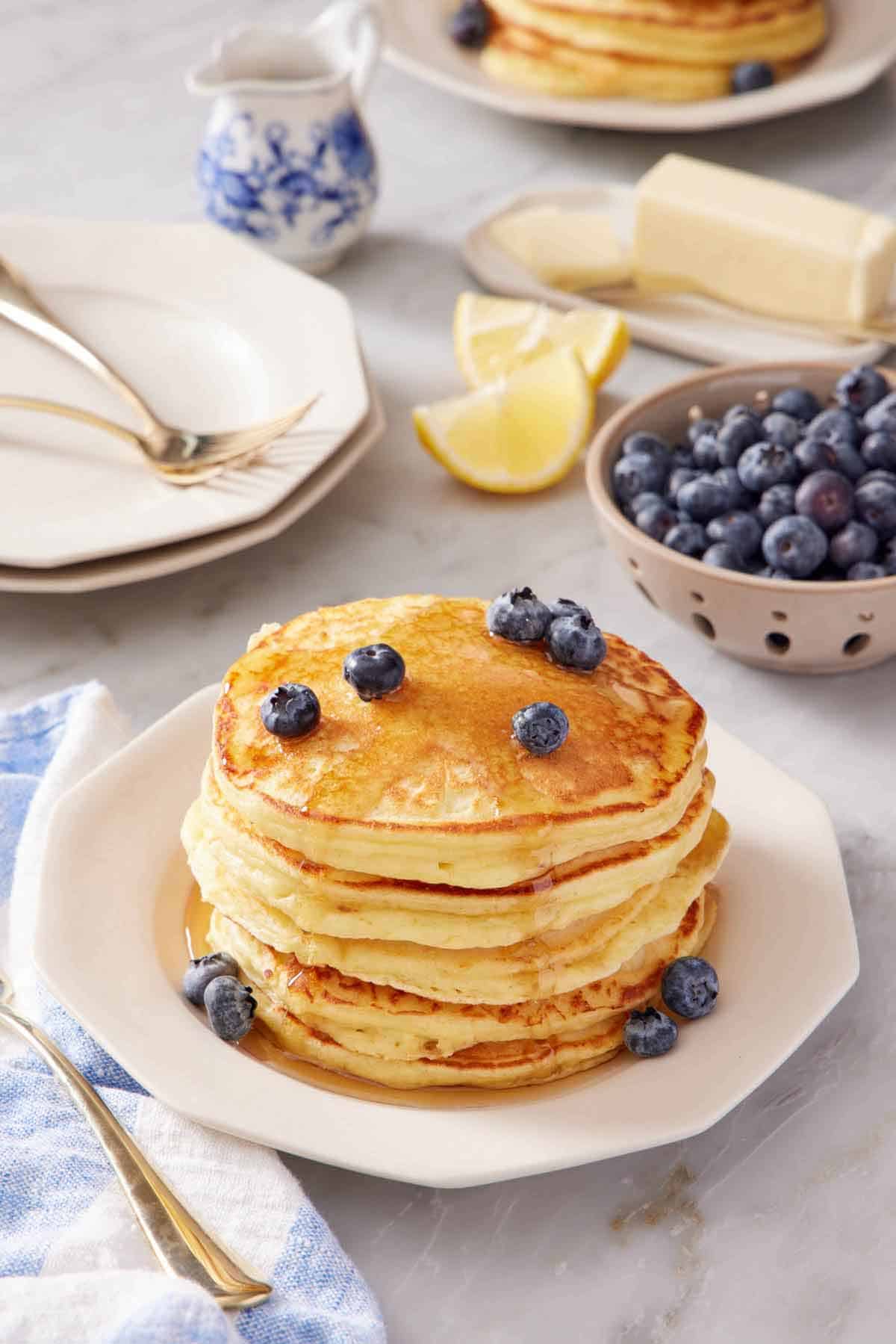 A stack of lemon ricotta pancakes topped with maple syrup and blueberries. A bowl of blueberries in the background along with plates, forks, lemon wedges, butter, and a small jug of syrup.