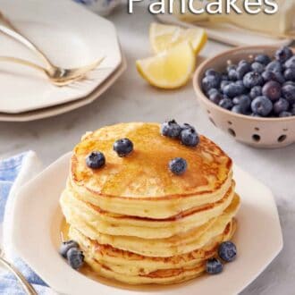 Pinterest graphic of a stack of lemon ricotta pancakes topped with maple syrup and blueberries. A bowl of blueberries in the background along with plates, forks, lemon wedges, butter, and a small jug of syrup.