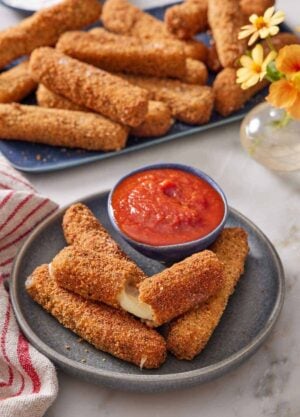 A plate with mozzarella sticks, one torn opened, with a bowl of marinara sauce. A platter of more mozzarella sticks in the background with a small vase of flowers.