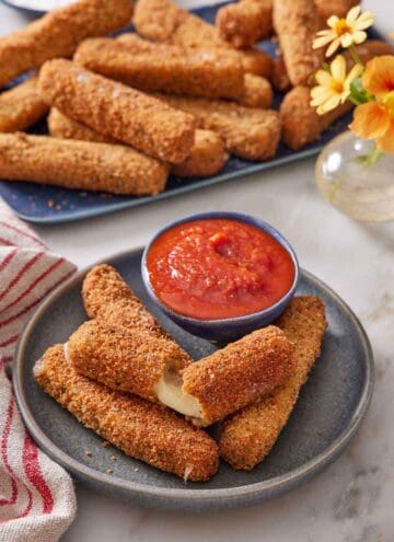 A plate with mozzarella sticks, one torn opened, with a bowl of marinara sauce. A platter of more mozzarella sticks in the background with a small vase of flowers.