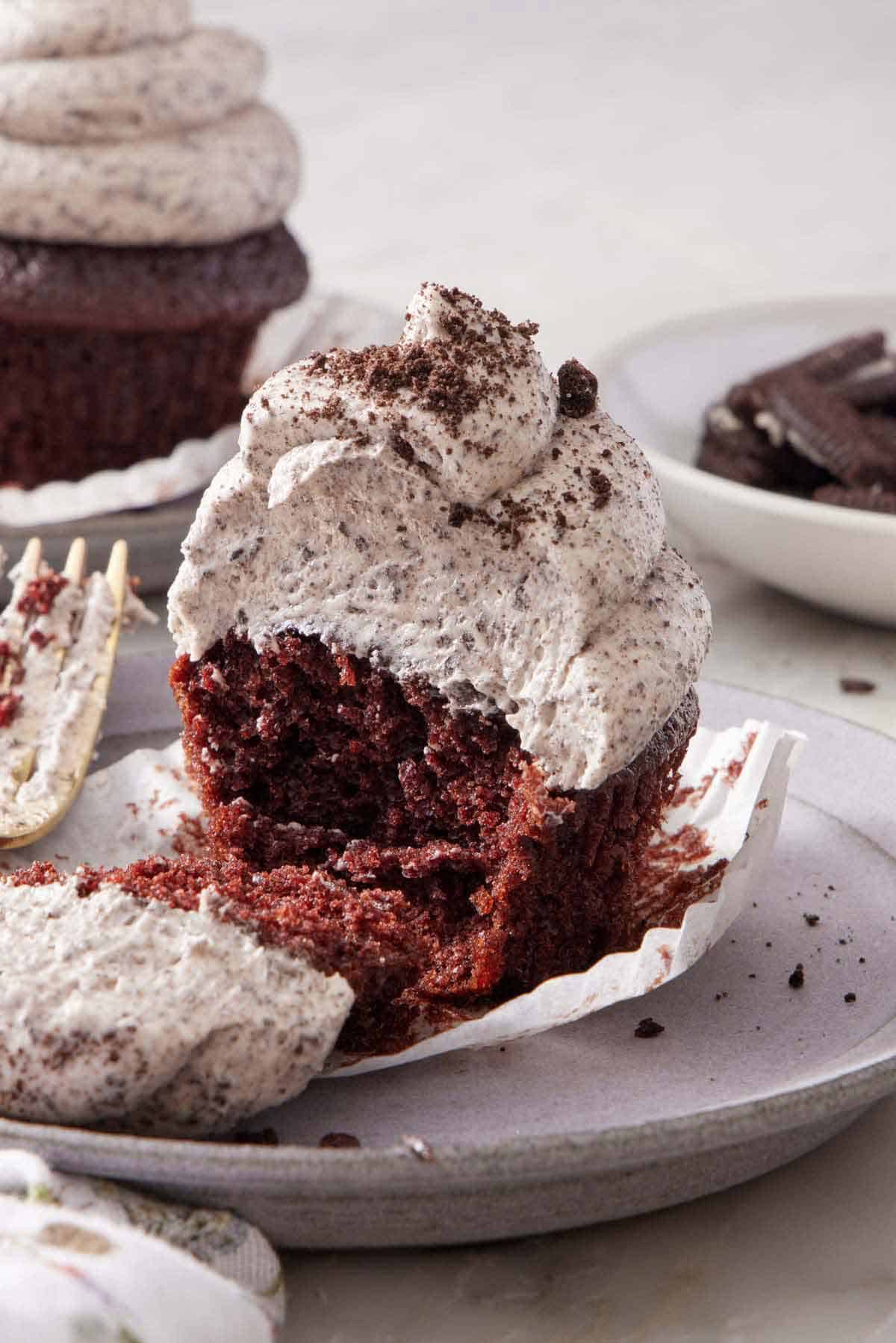 A chocolate cupcake topped with Oreo frosting, with a bite taken, showing the interior. Another frosted cupcake in the background with a bowl of Oreos.