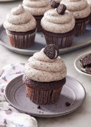 A cupcake topped with Oreo frosting and a mini oreo on a small plate. More Oreo frosted cupcakes in the background.