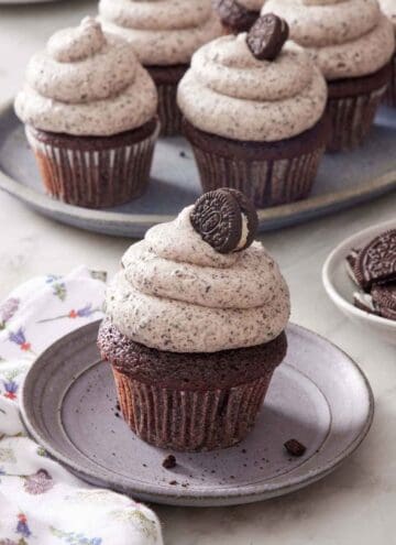 A cupcake topped with Oreo frosting and a mini oreo on a small plate. More Oreo frosted cupcakes in the background.