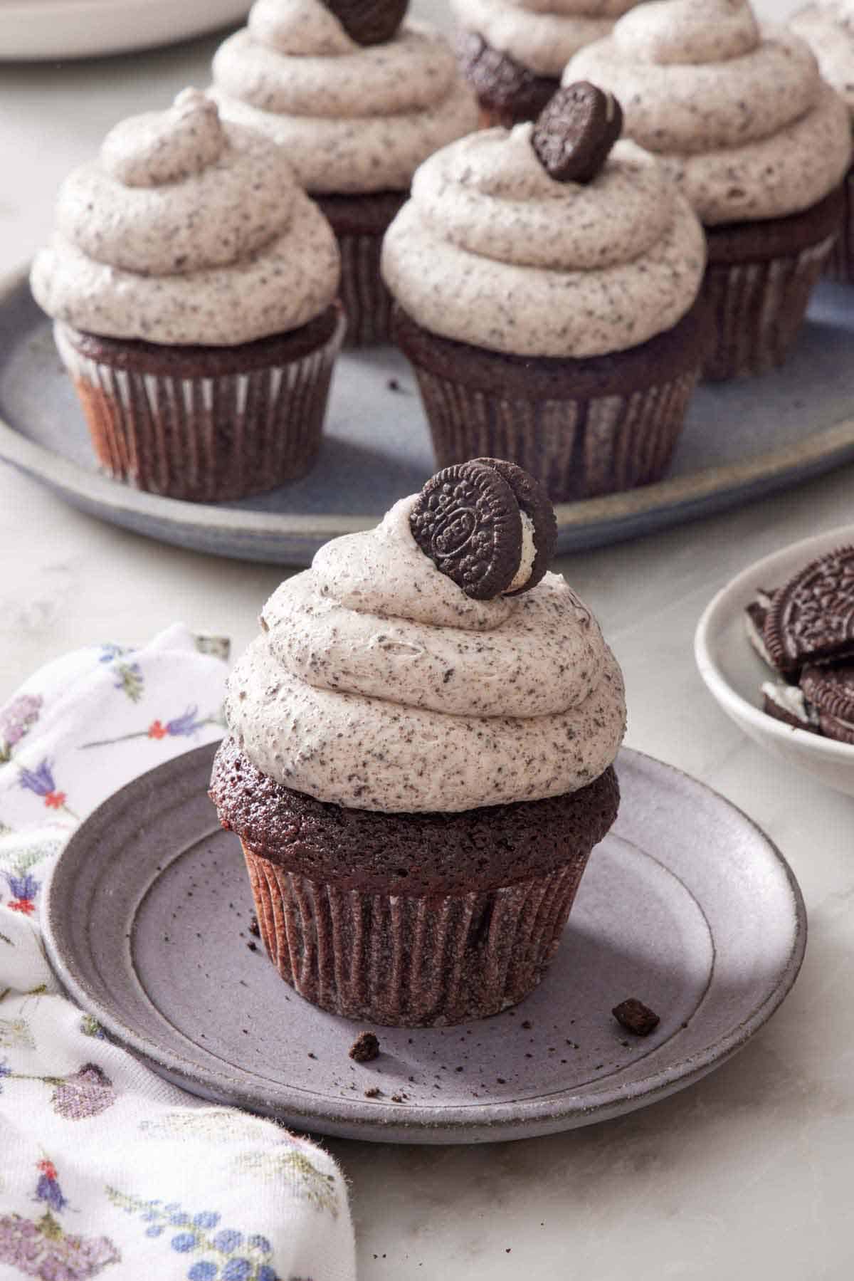 A cupcake topped with Oreo frosting and a mini oreo on a small plate. More Oreo frosted cupcakes in the background.